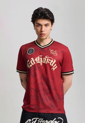 Junger Mann mit dunklen Haaren, der ein rotes Ed Hardy-Shirt mit schwarzen Drachendesigns, goldener Schrift und schwarz gestreiftem Kragen sowie Ärmelrändern trägt.