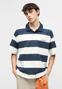 Νεαρός άνδρας με κοντά σκούρα μαλλιά, ντυμένος με μια μπλε και λευκή ριγέ πολο μπλούζα και κρατώντας ένα πορτοκαλί καπέλο, φορώντας μπεζ παντελόνι.