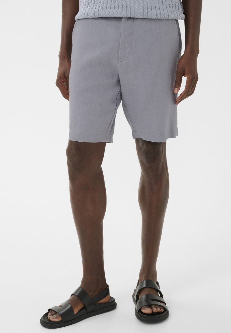 Homme portant un short gris clair et des sandales à brides noires, debout devant un fond blanc uni.