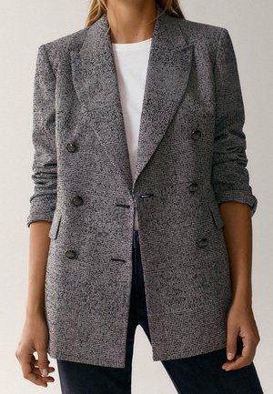 Kobieta nosząca szarą kratowaną dwurzędową marynarkę na białej koszuli, w połączeniu z ciemnymi spodniami, stoi z rozluźnionymi ramionami.