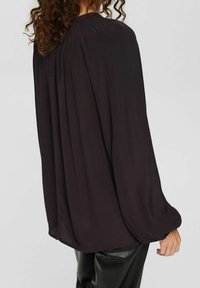 Blouse noire à manches longues avec une coupe décontractée, ornée de plis aux épaules, texture douce et subtil éclat. Associée à un pantalon noir.