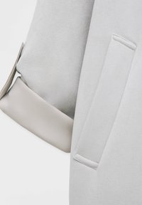 Manche d’un manteau en laine gris clair avec un poignet en cuir beige plié et une poche verticale sur le corps du manteau.