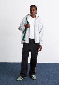 Gray zip-up hoodie, white t-shirt, black sweatpants, green shoes. Model carries a small cream bag with a green strap, standing on blue carpet.
