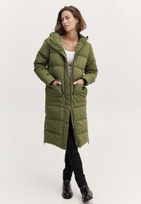Duga maslinasto zelena puffer jakna s kapuljačom, prednjim zatvaračem, bočnim džepovima i kviltanim dizajnom. Nosi se preko crnih hlača i čizmica do gležnja.