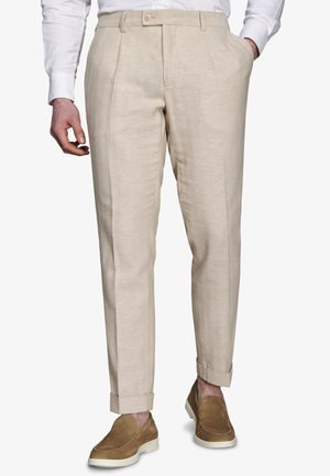 Beżowe dopasowane spodnie z plisami z przodu, noszone z brązowymi zamszowymi mokasynami i białą koszulą, ręce swobodnie ułożone.