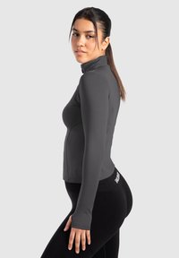 Grå langærmet sportsbluse med høj krave, lavet af glat materiale, tætsiddende design og tommelfingerhul ved manchetterne. Sorte leggings er synlige.