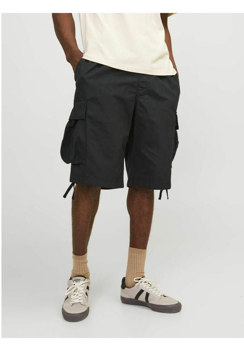 Schwarze Cargo-Shorts aus Stoff mit großen Seitentaschen, Kordelzugsäumen und lässiger Passform. Kombiniert mit grauen und schwarzen Sneakers sowie beigen Socken.