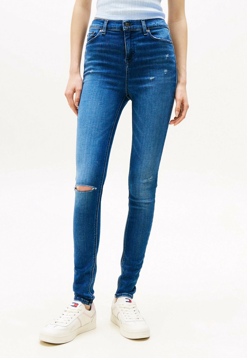 Smala blå jeans av denim, med en slitna design som har ett knähål och subtila blekningar, tillsammans med vita sneakers.