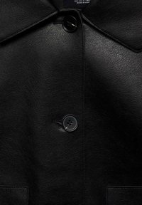 Sort læderjakke med en glat tekstur, der har en rund krave og en fremtrædende knaplukning. Inkluderer en brystlomme.