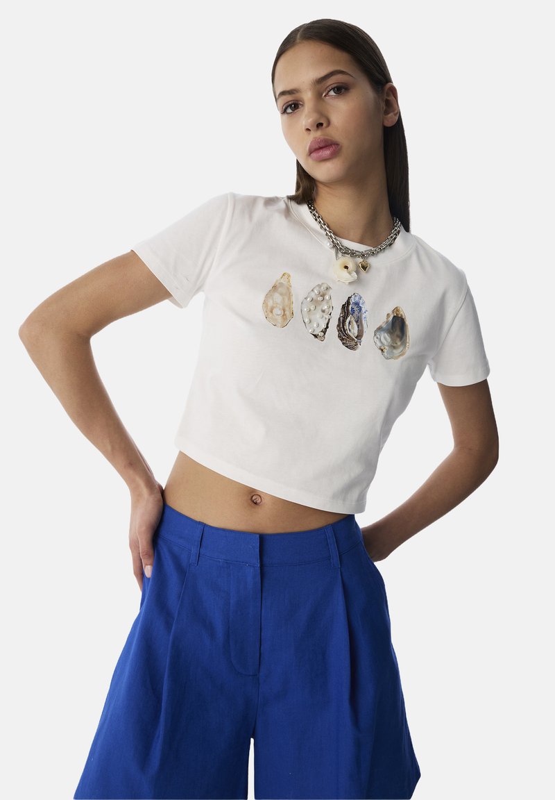 T-shirt blanc court avec cinq coquilles d'huître imprimées dans différentes couleurs et textures. Assorti à un short bleu ample.