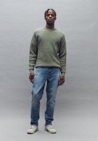 Suéter de canalé verde claro, jeans azules relajados y zapatillas blancas. El suéter tiene un escote redondo y mangas largas. Equilibra textura y simplicidad.