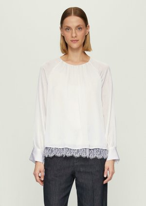 Blouse blanche à manches longues avec une texture plissée, col rond et bordure en dentelle à l'ourlet. Présente des poignets ajustés avec un léger évasement.