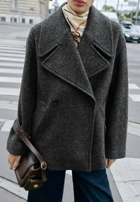 Cappotto doppiopetto in misto lana grigio scuro con revers a punta, tasche frontali e una vestibilità comoda, abbinato a una borsa a tracolla marrone.