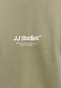 Ύφασμα ελιάς με λευκό κείμενο λογότυπο "JJ Studios" και μικρότερο κείμενο που αναγράφει "Παραγωγή από The J&J Crew, Σχεδιασμένο στη Δανία."