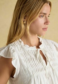 Weiße, leichte Bluse aus Stoff mit Rüschenschößen, einem gerafften Ausschnitt und einer Bindeschleife an der Vorderseite. Dezente Piqué-Textur im gesamten Design.