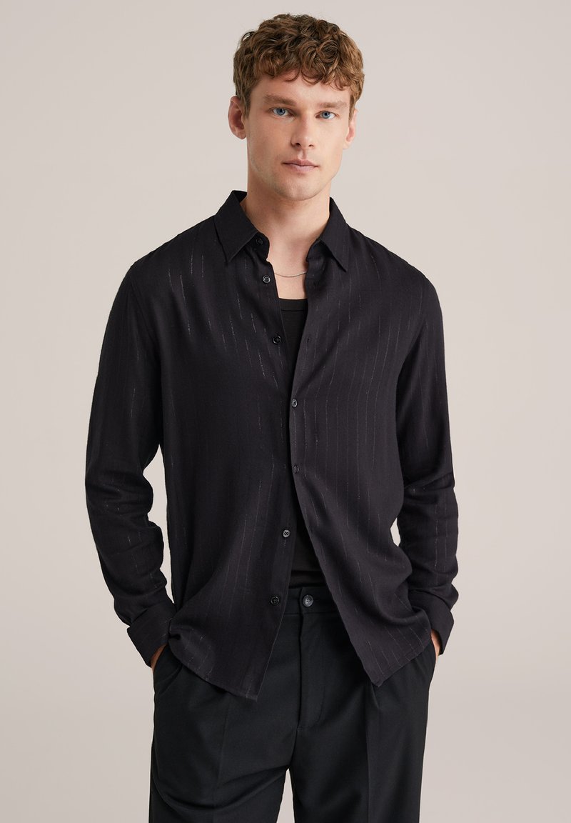 Mustast nööpidega särk, millel on peened vertikaalsed triibud, pikad varrukad, tekstureeritud kangas ja kraega kaelus, koos musta alussärgiga.