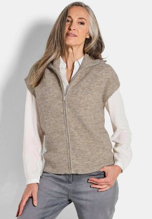 Gilet en tricot beige avec fermeture éclair et manches courtes, porté sur une chemise blanche. Présente un motif texturé et un ourlet arrondi.