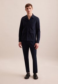 Giacca blu navy con due tasche anteriori e chiusura a pulsante, abbinata a pantaloni su misura dello stesso colore e scarpe eleganti nere. Texture liscia.