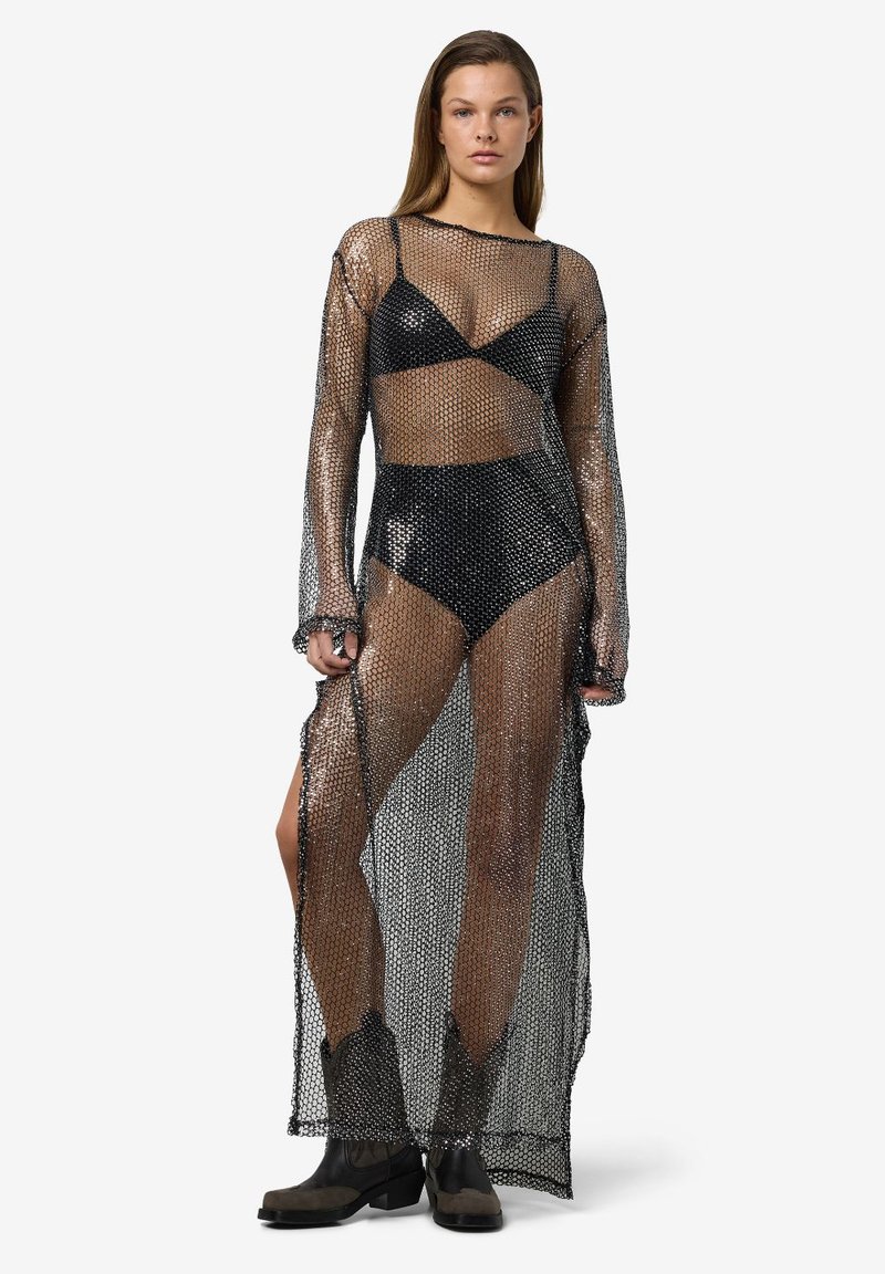 Vestido longo preto em malha com mangas longas, apresentando um design translúcido. Usado sobre um conjunto de biquíni preto correspondente. Comprimento até os tornozelos com aberturas laterais.