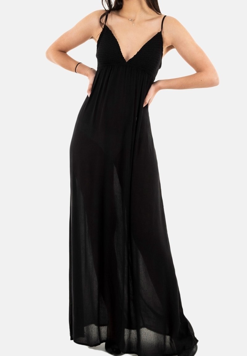 Femme portant une robe maxi noire sans manches avec de fines bretelles, debout les mains sur les hanches devant un fond uni.