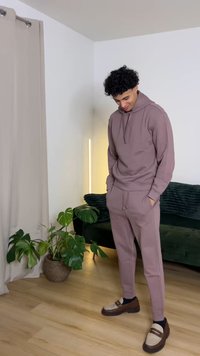 Hellvioletter Kapuzenpullover mit Kängurutasche, passende Hose und braune Loafers mit cremefarbenen Akzenten. Innenraum mit grünem Pflanzen und Sofa.