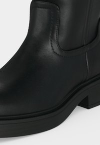 Bottine noire en cuir avec un bout arrondi, panneau latéral élastique et petit talon épais sur fond blanc.