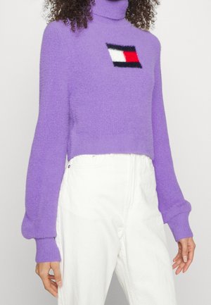 Femme portant un pull à col roulé violet en tissu duveteux avec un logo rectangulaire rouge, blanc et noir sur la poitrine, associé à un pantalon blanc taille haute.