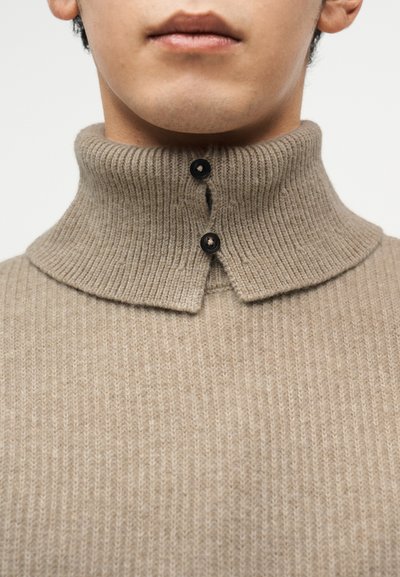 Μπεζ ριμπ πλεκτό πουλόβερ με ψηλό γιακά, διαθέτει κούμπωμα με κουμπιά κοντά στον λαιμό και υφασμάτινη υφή για επιπλέον λεπτομέρεια.