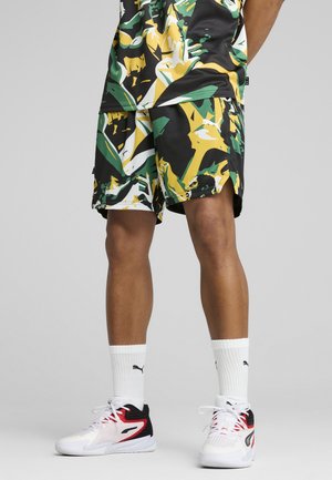 Jambes d'une personne portant un short camouflage vert, jaune et noir, des chaussettes Puma blanches et des baskets blanches, noires et rouges sur un sol gris.