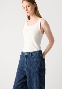 Valge ilma varrukateta tipp, mis on valmistatud elastsetest materjalidest, on kombineeritud kõrge talje ja lillemustriga siniste teksadega, millel on ka kuldne nööpide detail.