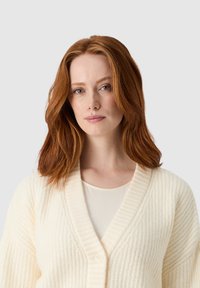 Kremowy sweter z dzianiny z dekoltem w serek, o prążkowanej strukturze i dużych guzikach z przodu, noszony na jasnej bluzce, podkreślający miękki, przytulny design.