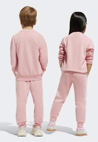 Svetloružové mikiny a teplákové nohavice s bielymi postrannými pruhmi a elastickými manžetami, nosené s svetlými teniskami.
