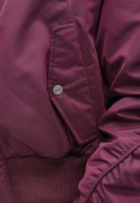 Vīna sarkanas krāsas polsterēta jaka ar ribotu apakšmali, sānu kabatu ar spiedpogām un savāktām piedurkņu detaļām, kas piešķir papildus tekstūru.