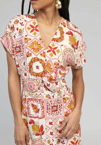 Robe à motifs avec un décolleté en V profond, des manches courtes et une taille cintrée. Présente des motifs floraux et géométriques en rouge, jaune et rose sur un fond crème.