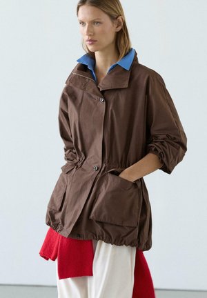 Frau mit hellbraunen Haaren trägt eine lockere braune Jacke über einem blaukragen Hemd, kombiniert mit weißen Hosen und einem roten Pullover, der um ihre Taille gebunden ist.