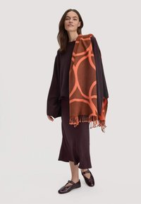 Vrouw in een donkerpaarse lange rok, oversized trui, zwarte schoenen en een oranje patroon sjaal over één schouder hangen, staande.