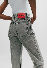 Harmaa farkud, joissa on korkea vyötärö ja suora leikkaus, kahdella takataskulla ja näkyvällä punaisella nahkalapulla, jossa lukee "HUGO", vyötärönauhassa.