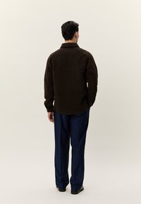 Vyras, stovintis nugara, dėvi tamsiai rudą ilgomis rankovėmis marškinius ir tamsiai mėlynas kelnes, priešais paprastą smėlio spalvos foną.