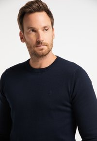 Tengerészkék kötött pulóver kerek nyakkivágással és bordázott mandzsettákkal, a mellel diszkrét horgonylogóval. Sima textúra.
