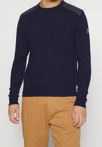 Tmavě modrý pletený svetr s dlouhými rukávy, žebrovaným lemem a detaily na ramenou z kontrastního materiálu. Na rukávu je umístěna logo záplata.
