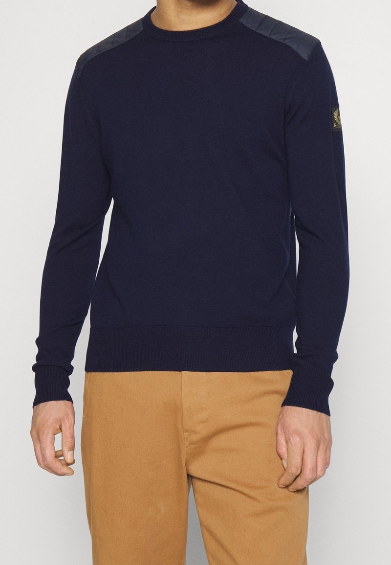 Tmavě modrý pletený svetr s dlouhými rukávy, žebrovaným lemem a detaily na ramenou z kontrastního materiálu. Na rukávu je umístěna logo záplata.