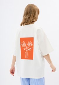Weißes übergroßes T-Shirt mit einem großen orangefarbenen Rechteck auf dem Rücken, das eine weiße Strichzeichnung von Blumen in einer Vase zeigt. Kurze Ärmel.