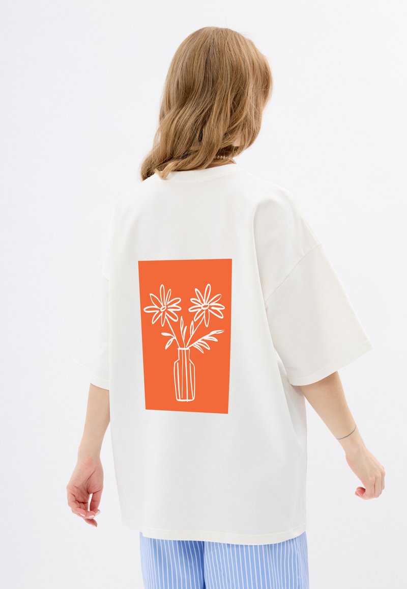 Witte oversized t-shirt met een grote oranje rechthoek op de achterkant, met een witte lijntekening van bloemen in een vaas. Korte mouwen.