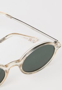 Kirkasta muoviset aurinkolasit, joissa on pyöreät tummat linssit ja ohut hopeametallinen varsi. Merkki "SEASALT" painettuna sivulle. Teksturoidut yksityiskohdat kehyksessä.