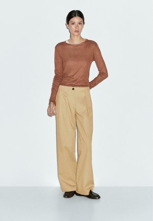 Woman wearing brown long-sleeve top, beige wide-leg pants, and black shoes standing against a white background.