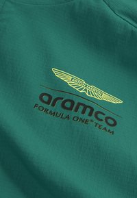 Tessuto verde con superficie strutturata. Testo nero "arameco" e logo alato giallo di "Aston Martin" ben visibili.