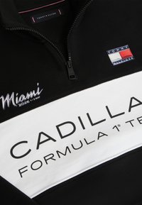 Cămașă alb-negru cu fermoar, cu textul „Cadillac Formula 1 Team”, detalii despre evenimentul Miami 2026 și o insignă cu logo-ul Tommy Hilfiger.