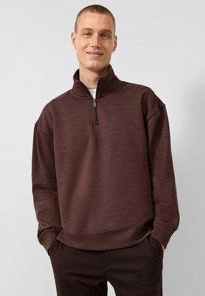 Man with short hair wearing brown half-zip sweatshirt and dark pants, standing with hands in pockets against a plain background.