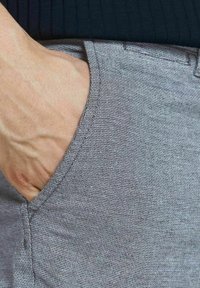 Szary, teksturowany materiał spodni, zbliżenie na okolicę kieszeni, z delikatnym wzorem plecionki i widoczną ręką opartą w kieszeni.