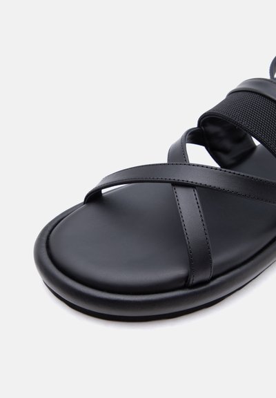 Μαύρο δερμάτινο σανδάλι με σταυρωτούς ιμάντες πάνω από τα δάχτυλα και φαρδύ ελαστικό ιμάντα κοντά στον αστράγαλο σε λευκό φόντο.